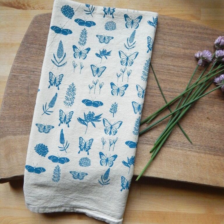 A hand-printed organic cotton tea towel, features a repeating pattern of blue butterflies and various blue botanical elements.