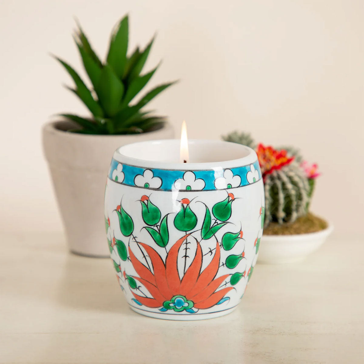 Decorative candle with floral design in a ceramic holder, surrounded by plants on a neutral background