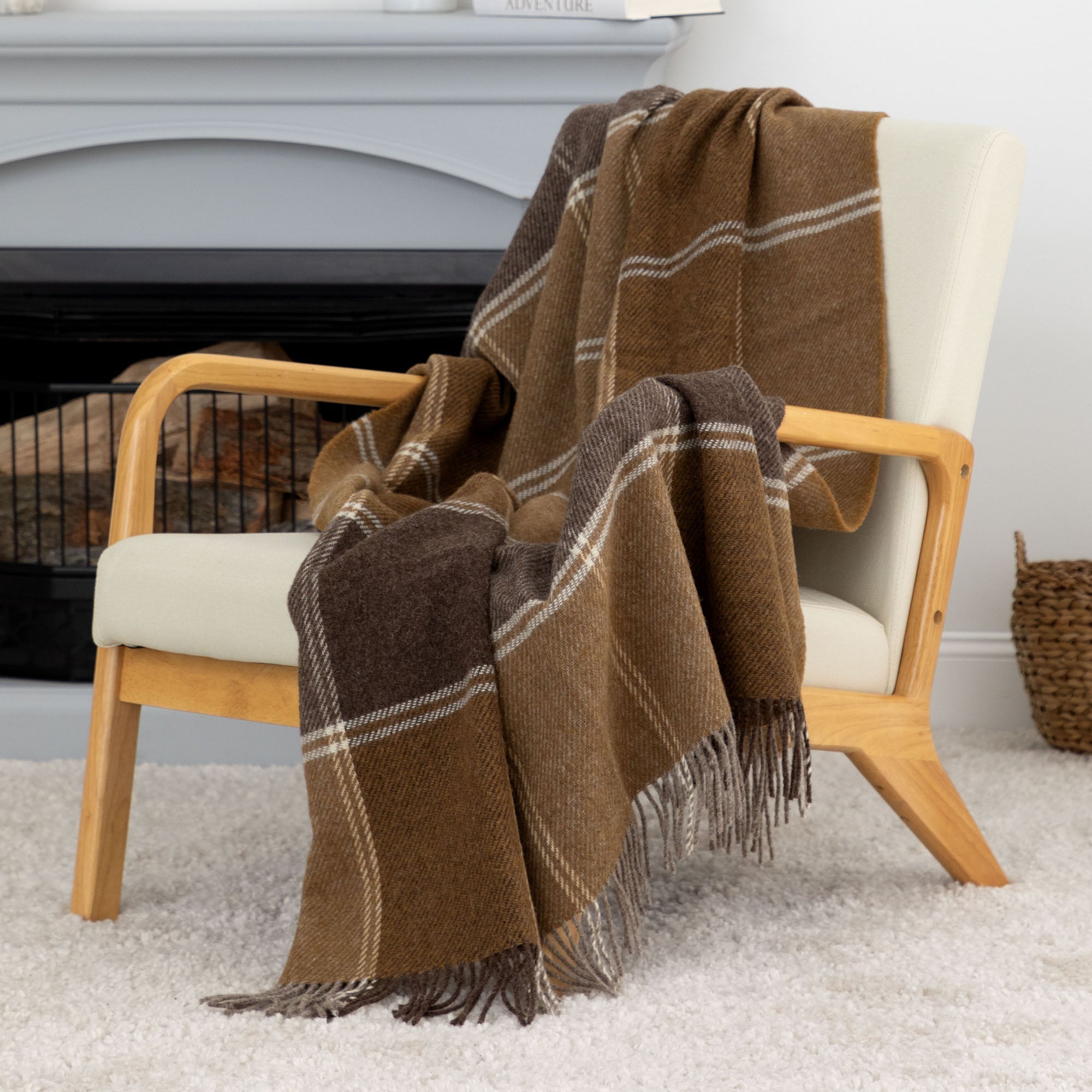 Brown plaid 100% wool throw blanket with fringe, draped over a modern cream and wood accent chair near a fireplace. Handmade in Ukraine.