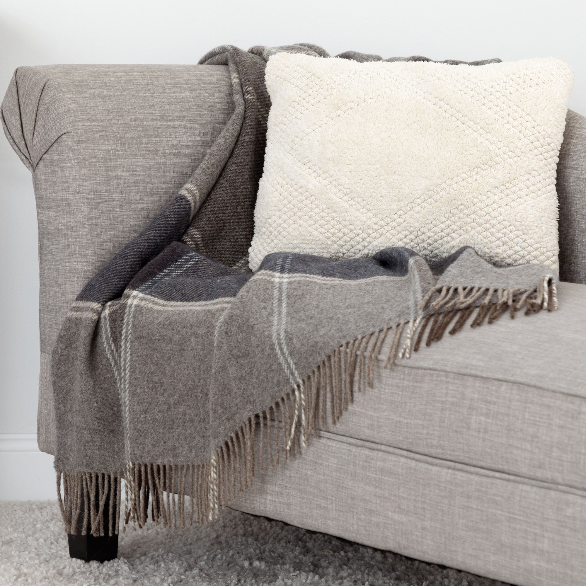 Gray and brown plaid wool throw with fringe draped over the arm of a beige sofa, accompanied by a textured cream throw pillow.