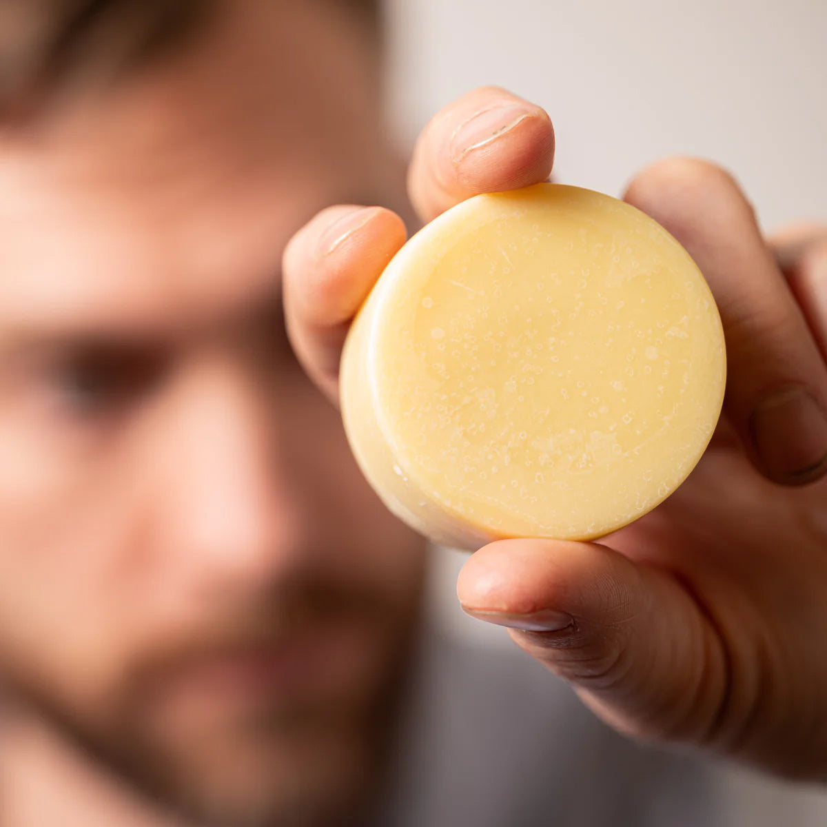 Hand holding a round Deep Hydration Cocoa Butter Lotion Bar with a blurred person in the background.