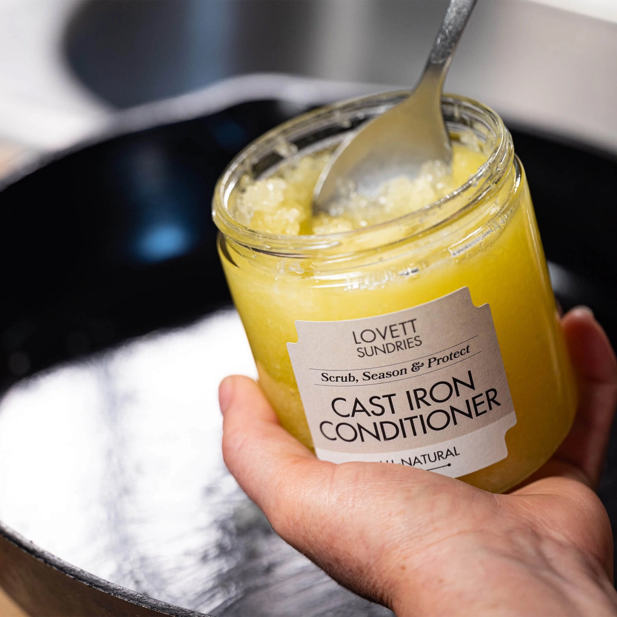 A hand holds a jar of "Lovett Sundries Cast Iron Conditioner" with a spoon inside. The yellow substance is held above a black cast iron pan.