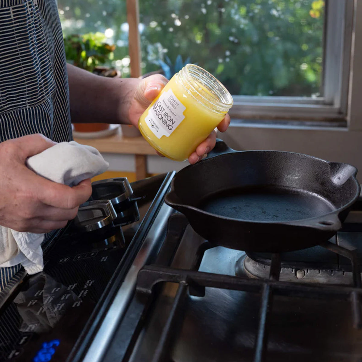 A person in a striped apron holds a jar of "Lovett Sundries Cast Iron Seasoning" in one hand and a white towel in the other. They are preparing to apply the seasoning to a cast iron pan on a kitchen stovetop.