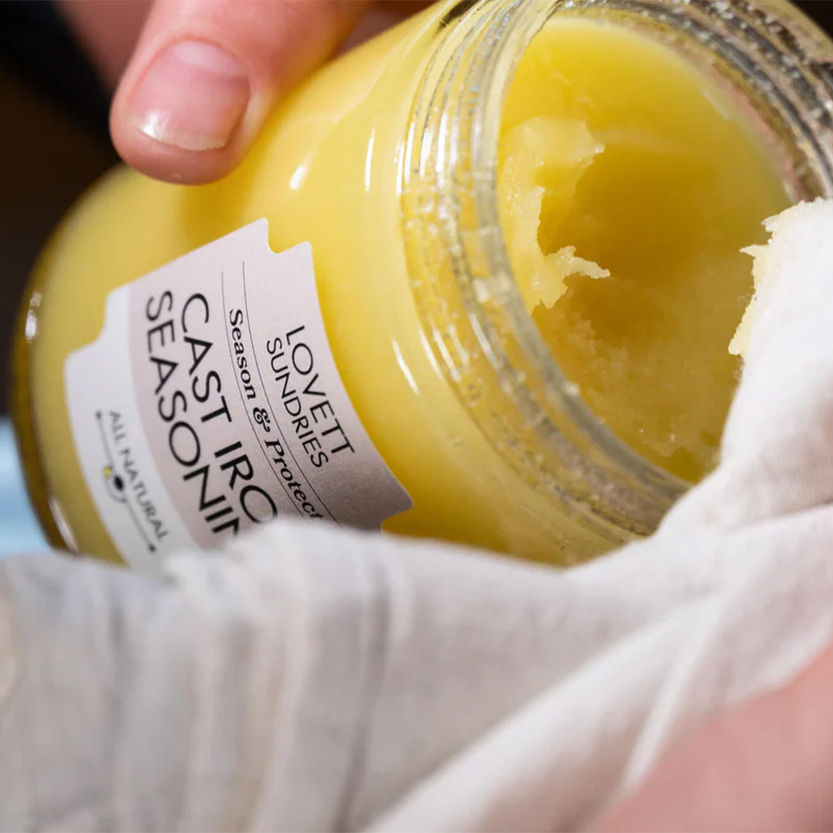 A hand holds a jar of "Lovett Sundries Cast Iron Seasoning" while wiping the side with a white cloth. A scoop has been taken from the yellow contents.
