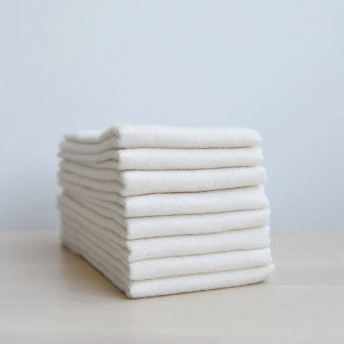A stack of multiple folded off-white towels is shown on a light-colored surface. The towels are stacked neatly and have no visible branding.