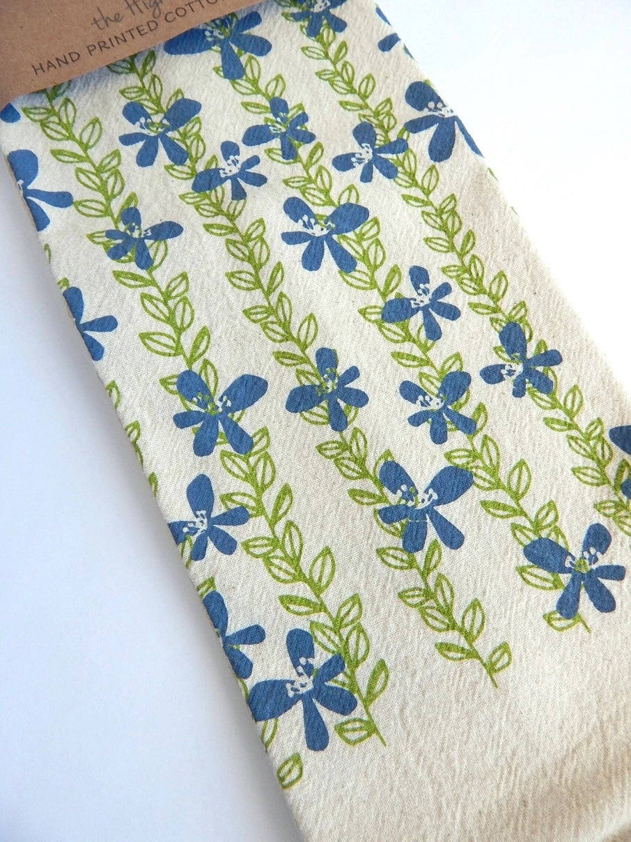 A close-up of a white hand-printed cotton tea cloth featuring vertical rows of blue flowers with green leafy stems.