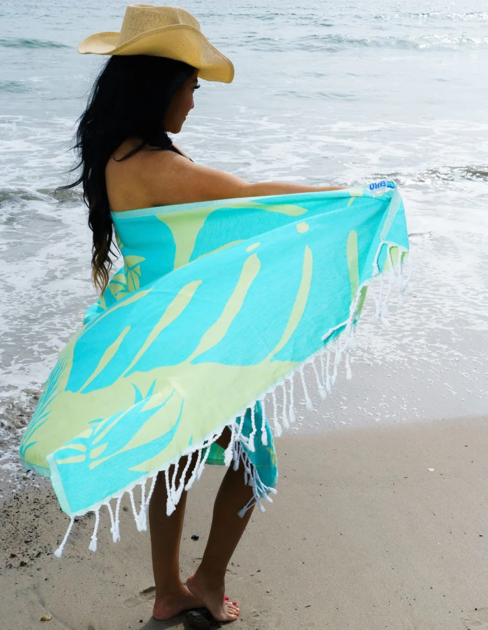 Woman in a sun hat holding a beach towel on a sunny beach, with the ocean in the background.