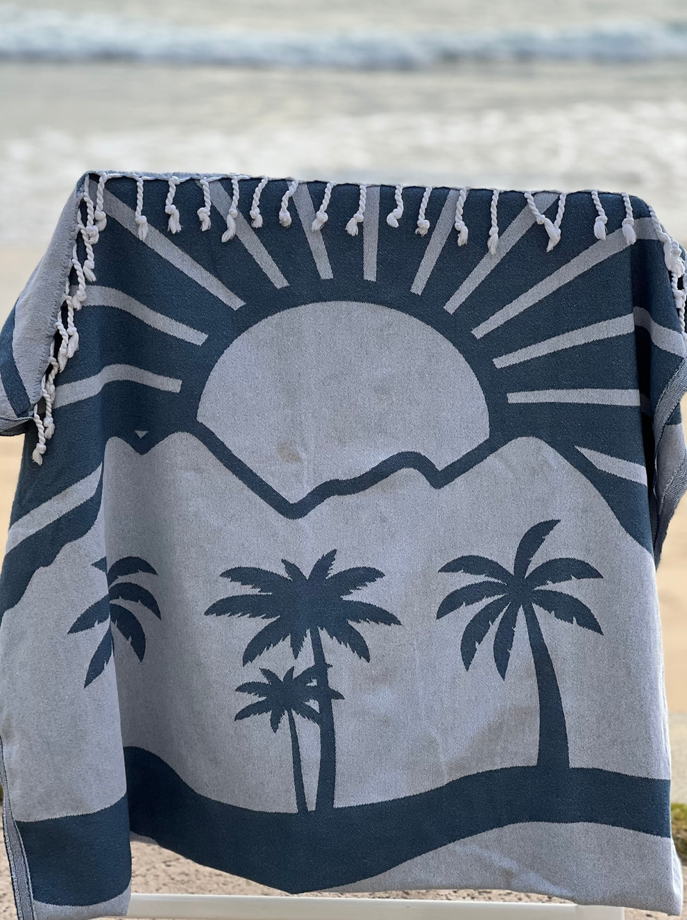 Turkish towel featuring a sunset, mountains, and beach design, hanging from a pole with the beach visible behind it.