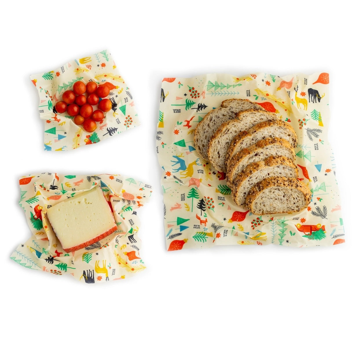 A flat lay photo showing reusable beeswax wraps with a festive forest animal pattern, containing sliced bread, a block of cheese, and cherry tomatoes on a white background.
