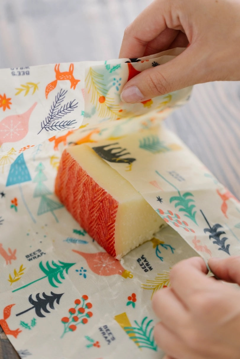 Hands wrapping cheese with beeswax food wrap featuring woodland animals and greenery on a cream background.