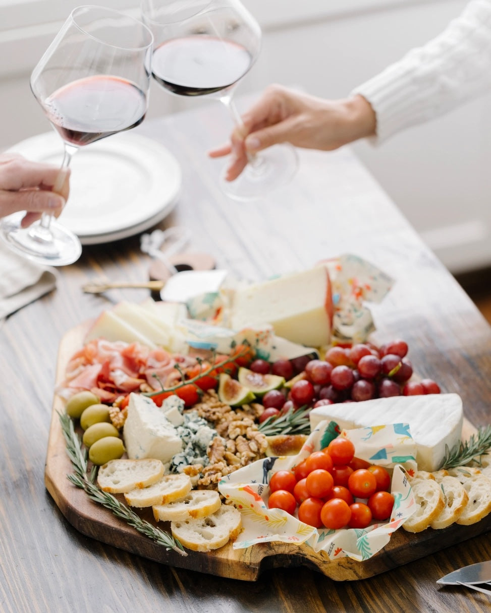 Close-up of a cheese board with cheeses, bread, grapes, figs, walnuts, cherry tomatoes in beeswax wraps, and two people toasting wine glasses.