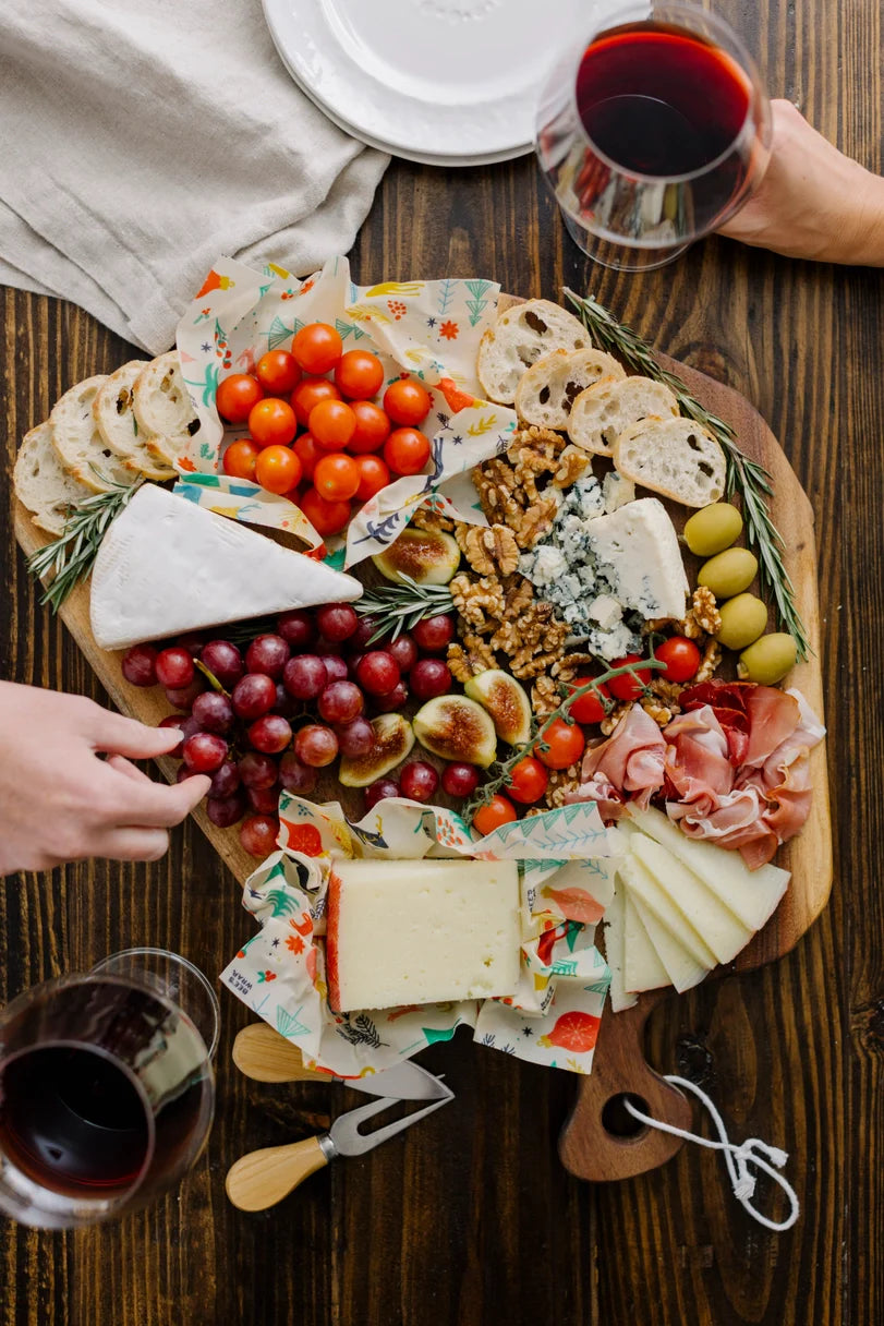 Overhead view of a cheese board with cheeses, grapes, figs, cherry tomatoes in beeswax wraps, walnuts, olives, sliced bread, and prosciutto with wine.