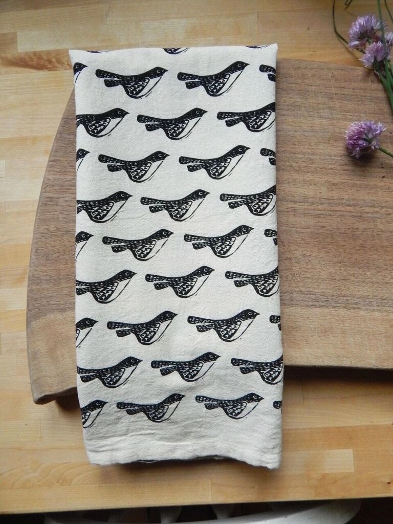 A hand-printed organic cotton tea towel, features a repeating pattern of stylized black birds.