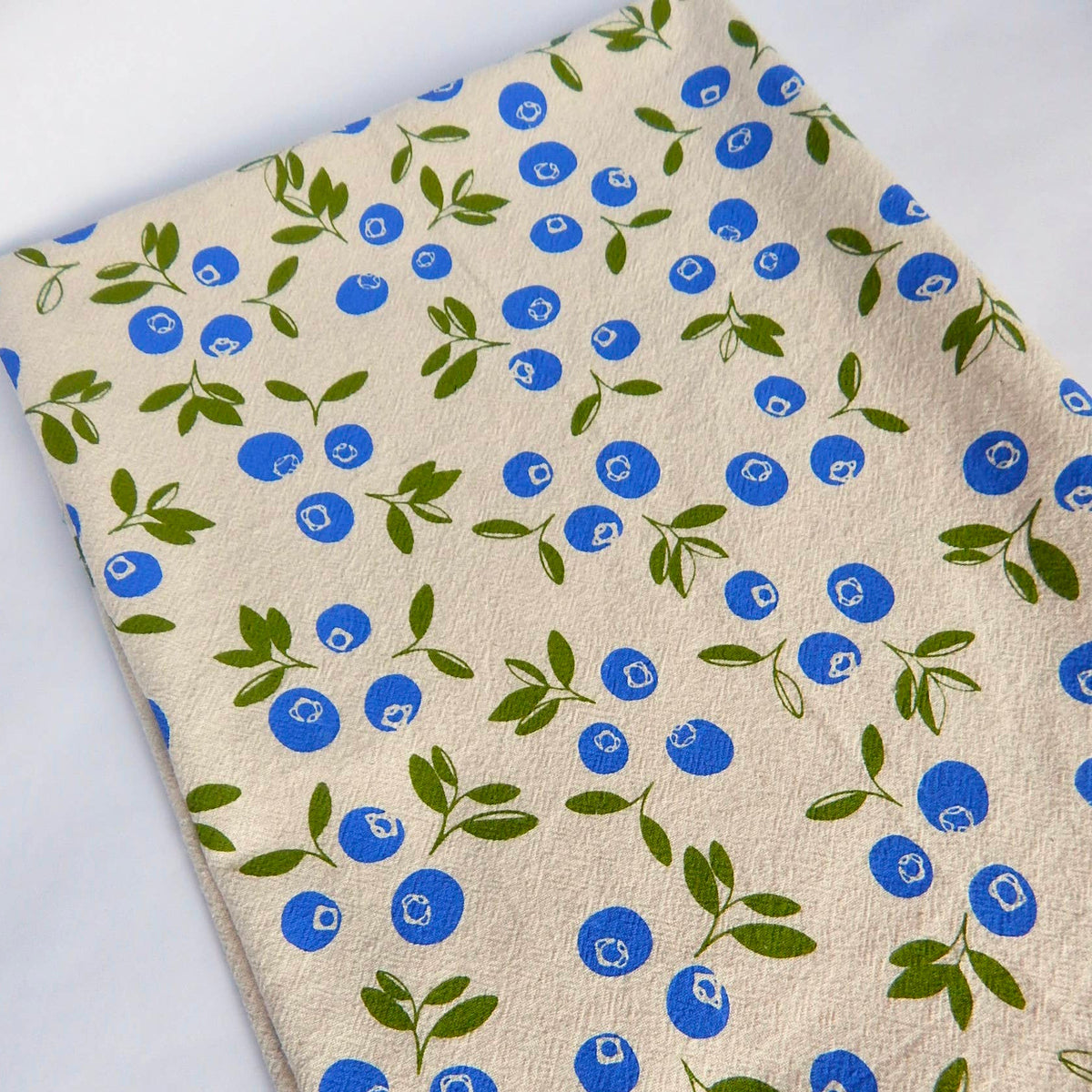 A hand-printed organic cotton tea towel, folded on a light surface, features a scattered pattern of bright blue blueberries and green leaves.