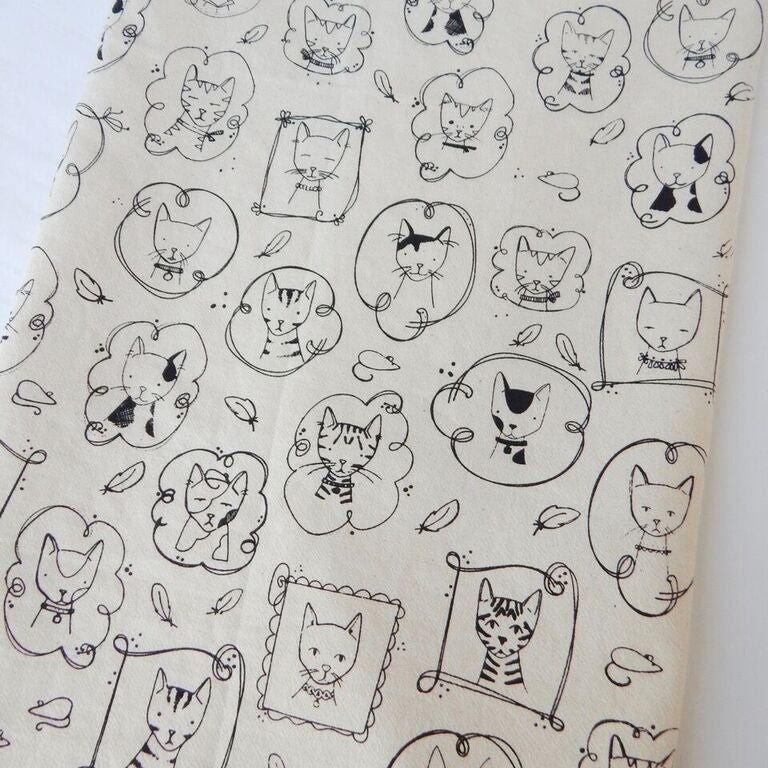 A hand-printed organic cotton tea towel, features a whimsical pattern of outlined black cats in various poses and frames, along with small feathers and mice prints.