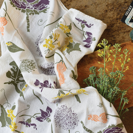 A white kitchen towel with a colorful botanical pattern of purple, yellow and peach flowers is draped over a wooden board with a bundle of green plants alongside it.