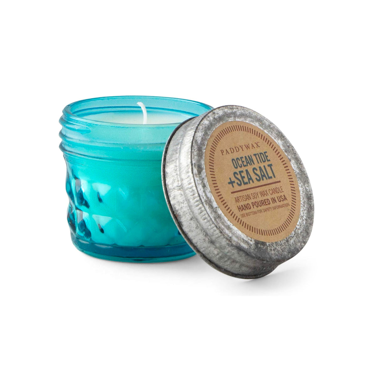 A small blue Paddywax artisan soy wax candle is shown in a jar with a textured diamond pattern. The lid is off, revealing a lit wick. The lid's label identifies the scent as Ocean Tide + Sea Salt.