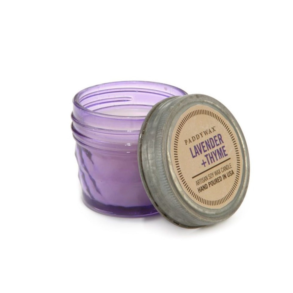 A small, purple Paddywax artisan soy wax candle is shown in a jar with the lid off to the side. The lid's label identifies the scent as Lavender + Thyme and notes it is hand-poured in the USA