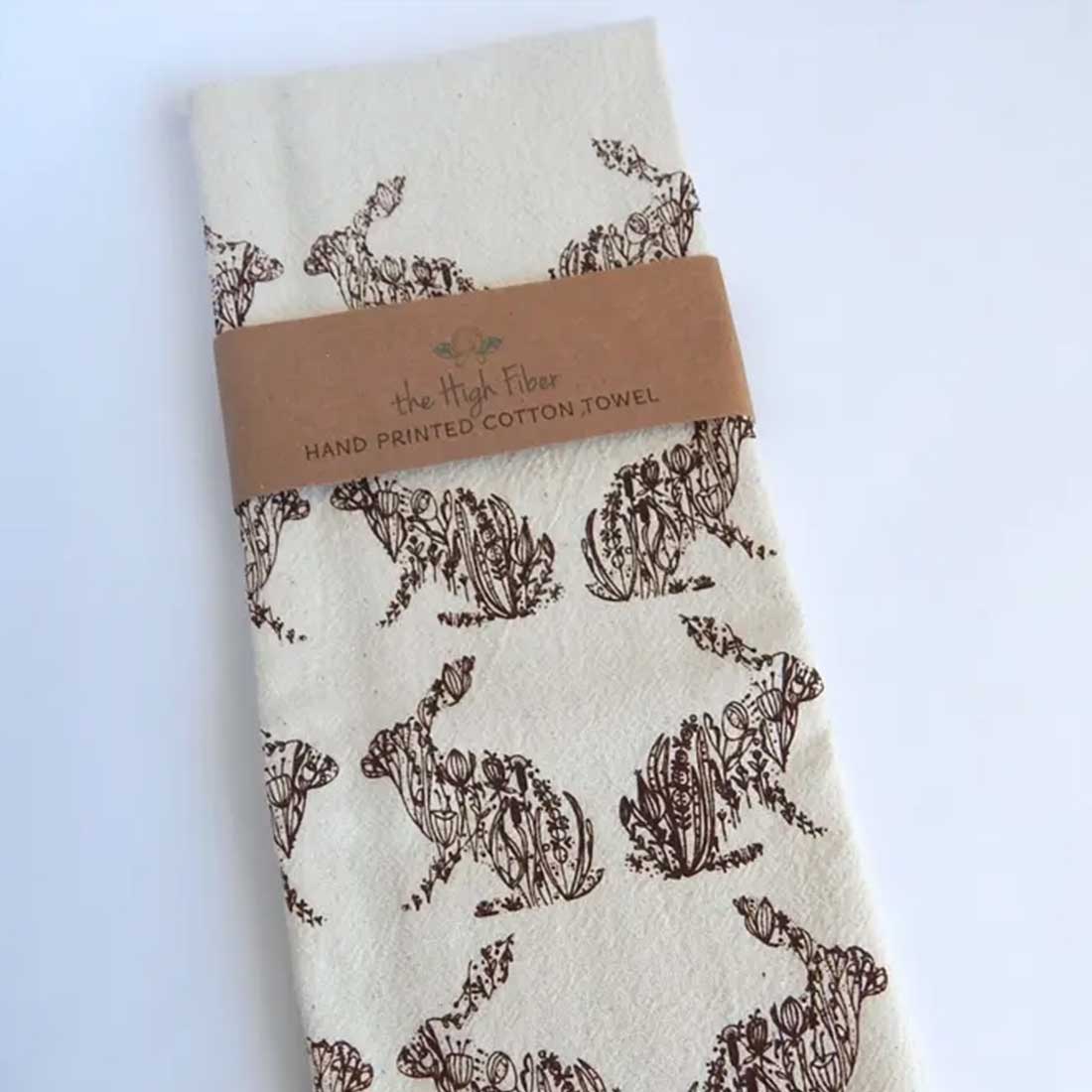 A hand-printed organic cotton tea towel from "the High Fiber," folded with a brown band. It features a repeating pattern of brown rabbits filled with intricate floral designs.